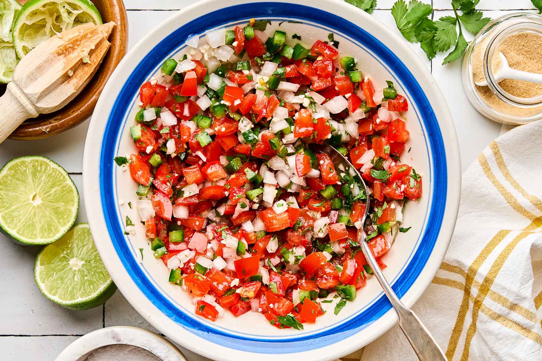 An overhead shot of fresh tomato salsa in a blue-rimmed white bowl atop a white tiled surface. Halved and juiced limes, dishes of garlic powder and salt, and a white and yellow plaid cloth napkin sit alongside the bowl.