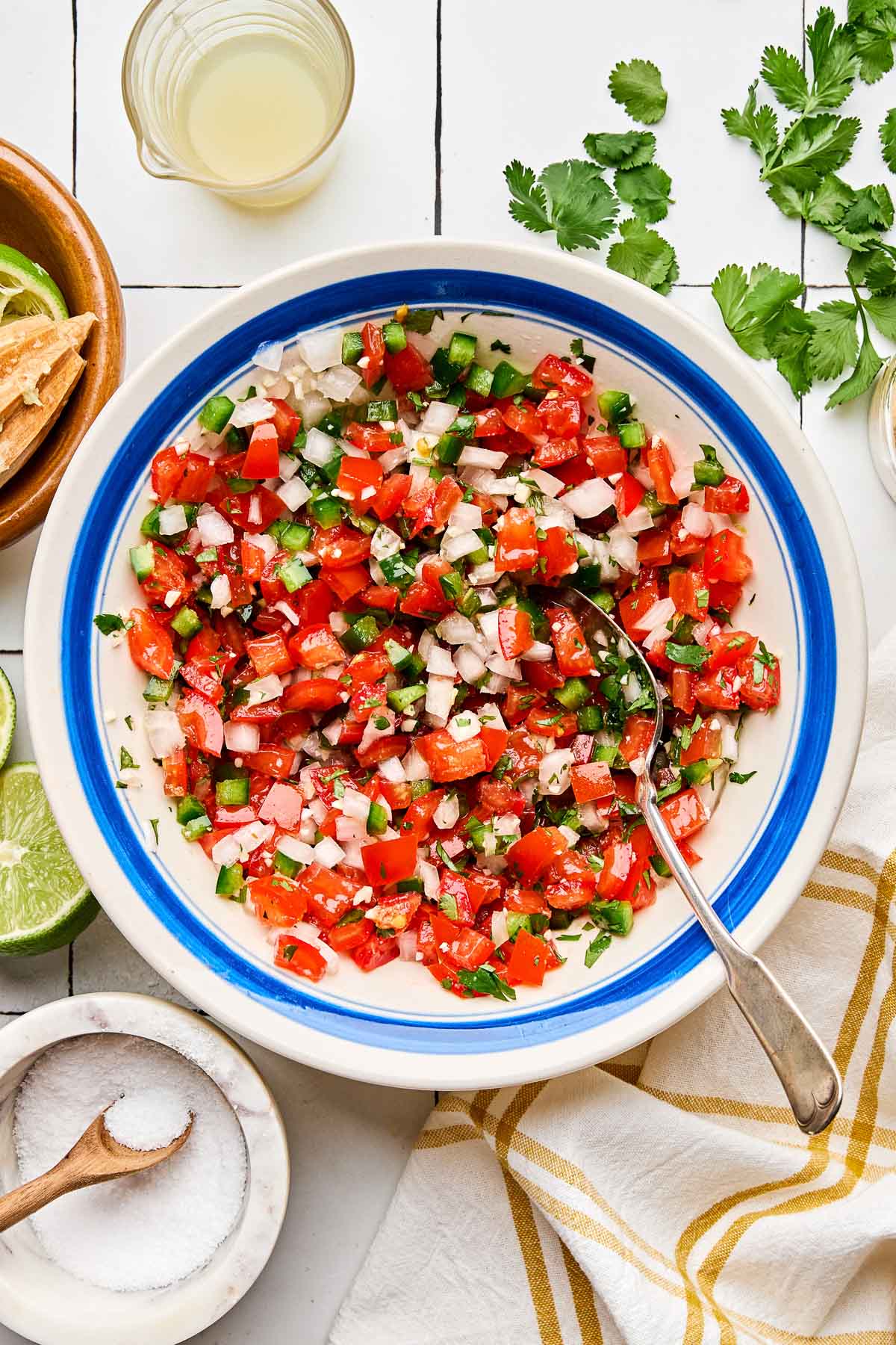 An overhead shot of fresh tomato salsa in a blue-rimmed white bowl atop a white tiled surface. Halved and juiced limes, cilantro, dishes of garlic powder and salt, and a white and yellow plaid cloth napkin sit alongside the bowl.