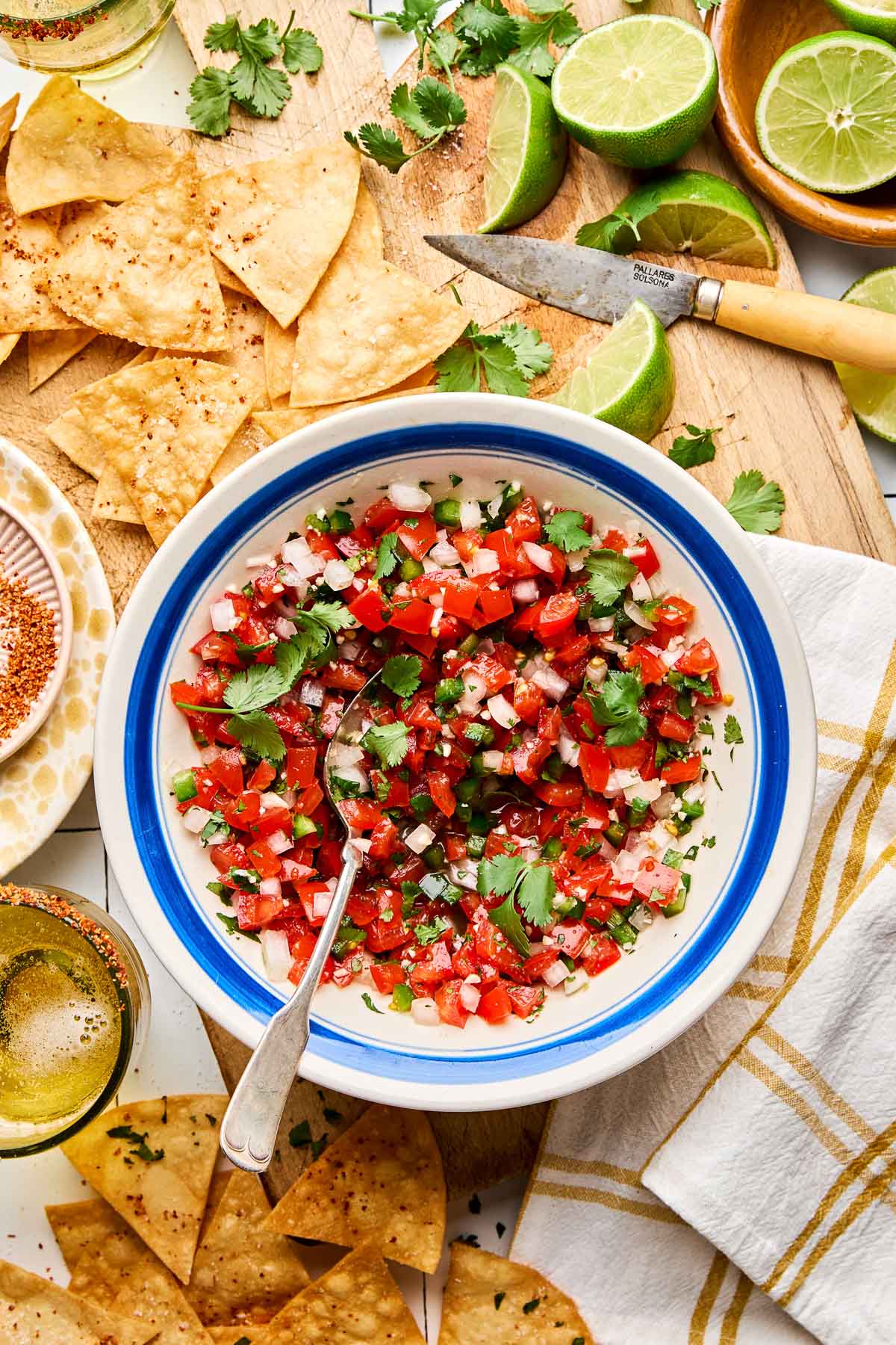An overhead shot of a bowl of fresh tomato salsa on a wooden cutting board atop a white tiled surface, surrounded by tortilla chips, lime wedges and halved limes, cilantro, a dish of tajin, and 2 margaritas.