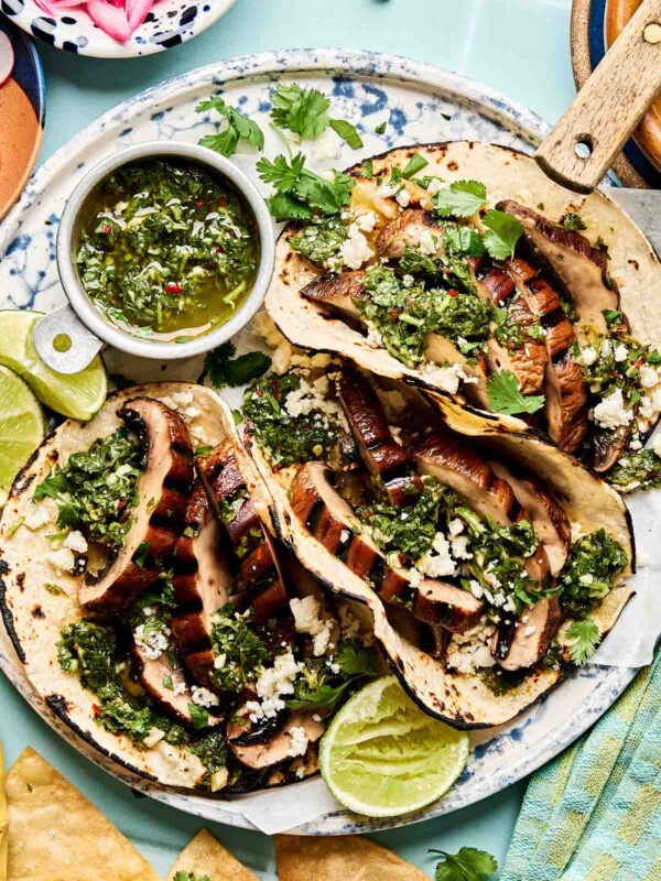 Three grilled portobello mushroom tacos on corn tortillas, topped with chimichurri sauce, crumbled cheese, and fresh cilantro, served with lime wedges and a small bowl of chimichurri on a patterned plate.