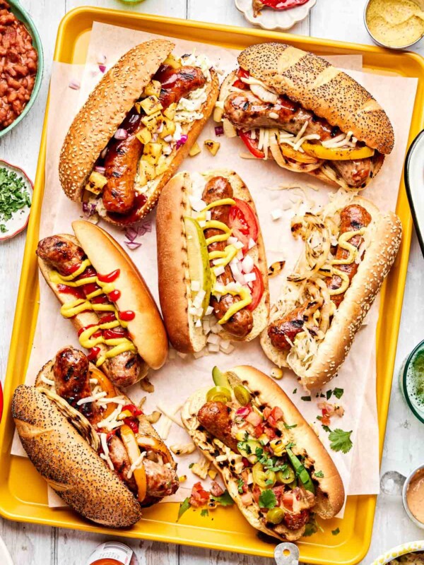 A tray with six assorted grilled sausages on buns, each topped with various ingredients like onions, tomatoes, pickles, relish, mustard, ketchup, and shredded cheese, surrounded by bowls of sauces and toppings on a light surface.