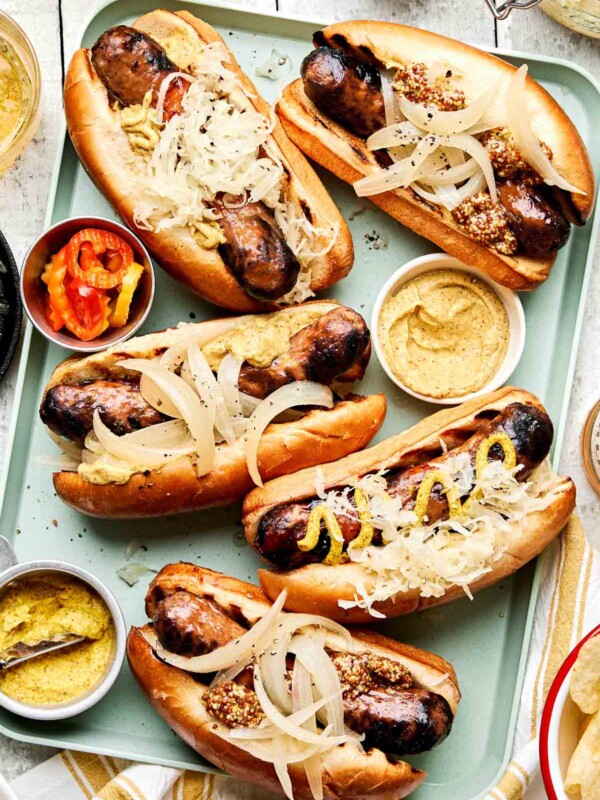 An overhead shot of 5 beer brats on brioche buns on a light green tray atop a white planked surface. The brats are topped with various condiments and onions.