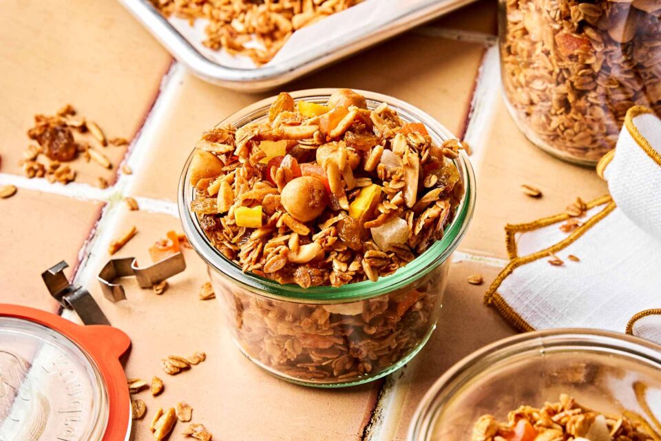 An angled shot of a glass bowl full of tropical fruit granola on a peach tiled surface. The bowl is surrounded by extra granola in another bowl, a storage canister, and on a parchment-lined baking sheet.