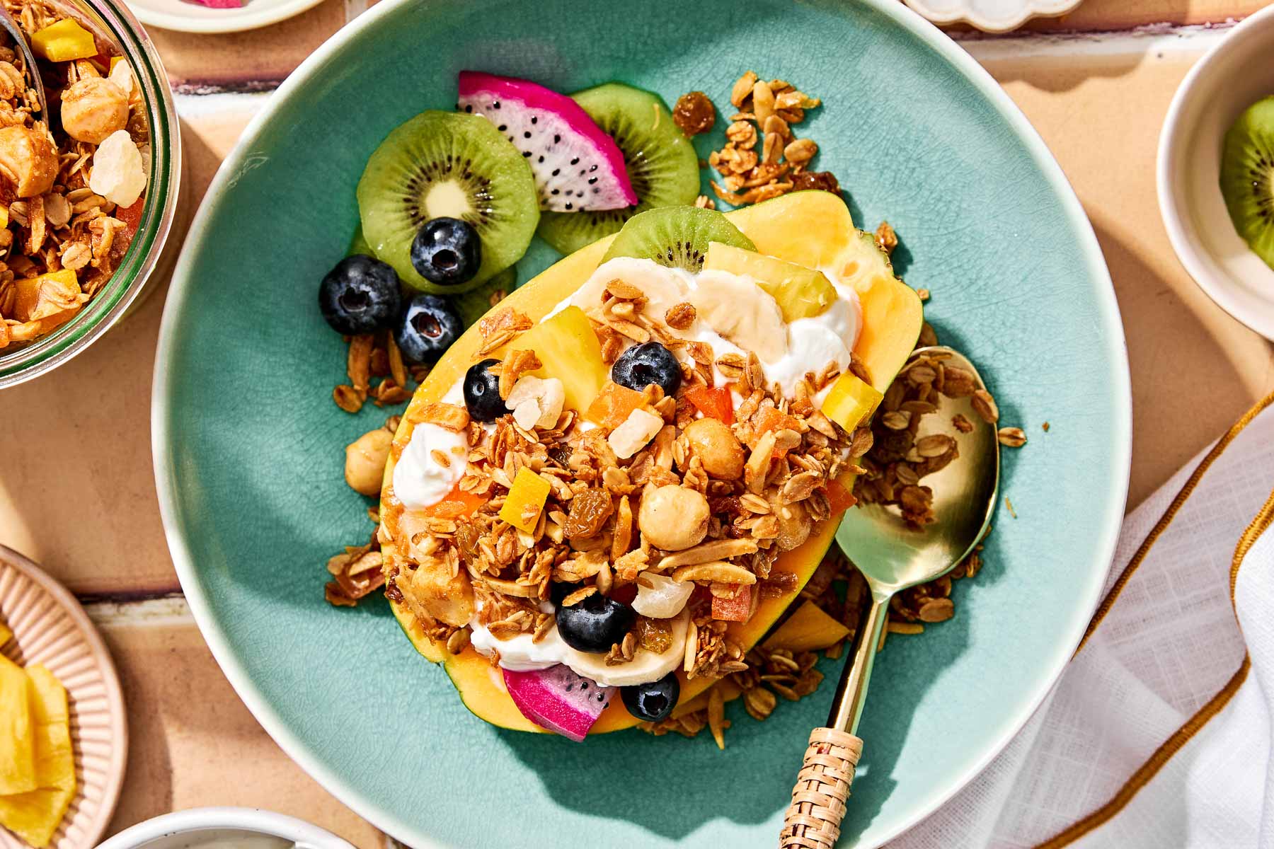 An overhead shot of a halved papaya topped with yogurt, fresh fruit and tropical granola. The papaya sits in a bright teal bowl on a peach-colored surface. A container of granola sits alongside the bowl.