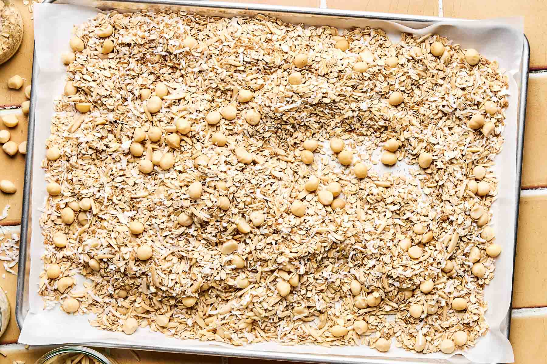 An overhead shot of dry ingredients - macadamia nuts, oats, almonds, coconut, and brown sugar - on a large rimmed baking sheet lined with parchment paper. The baking sheet sits on a peach tiled surface.