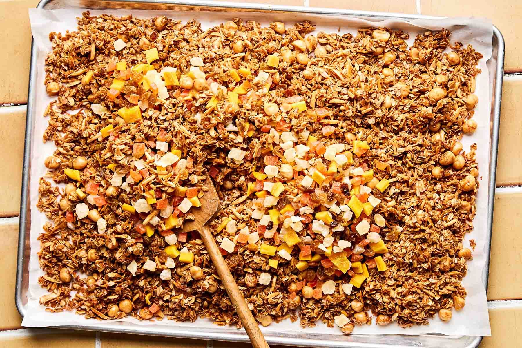 An overhead shot of a wooden spoon stirring diced dried fruit into baked granola on a parchment-lined baking sheet. The baking sheet sits on a peach-colored tiled surface.