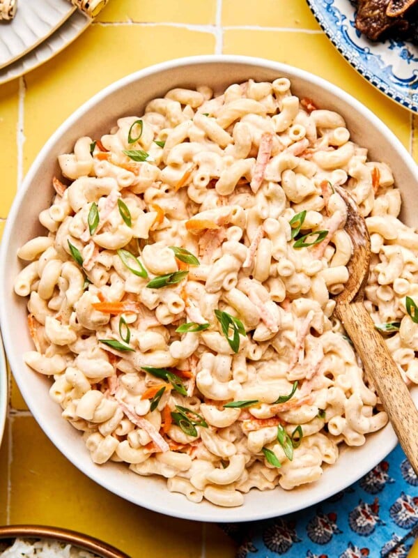 A bowl of creamy macaroni salad with sliced green onions and grated carrots, served with a wooden spoon on a yellow tiled surface.