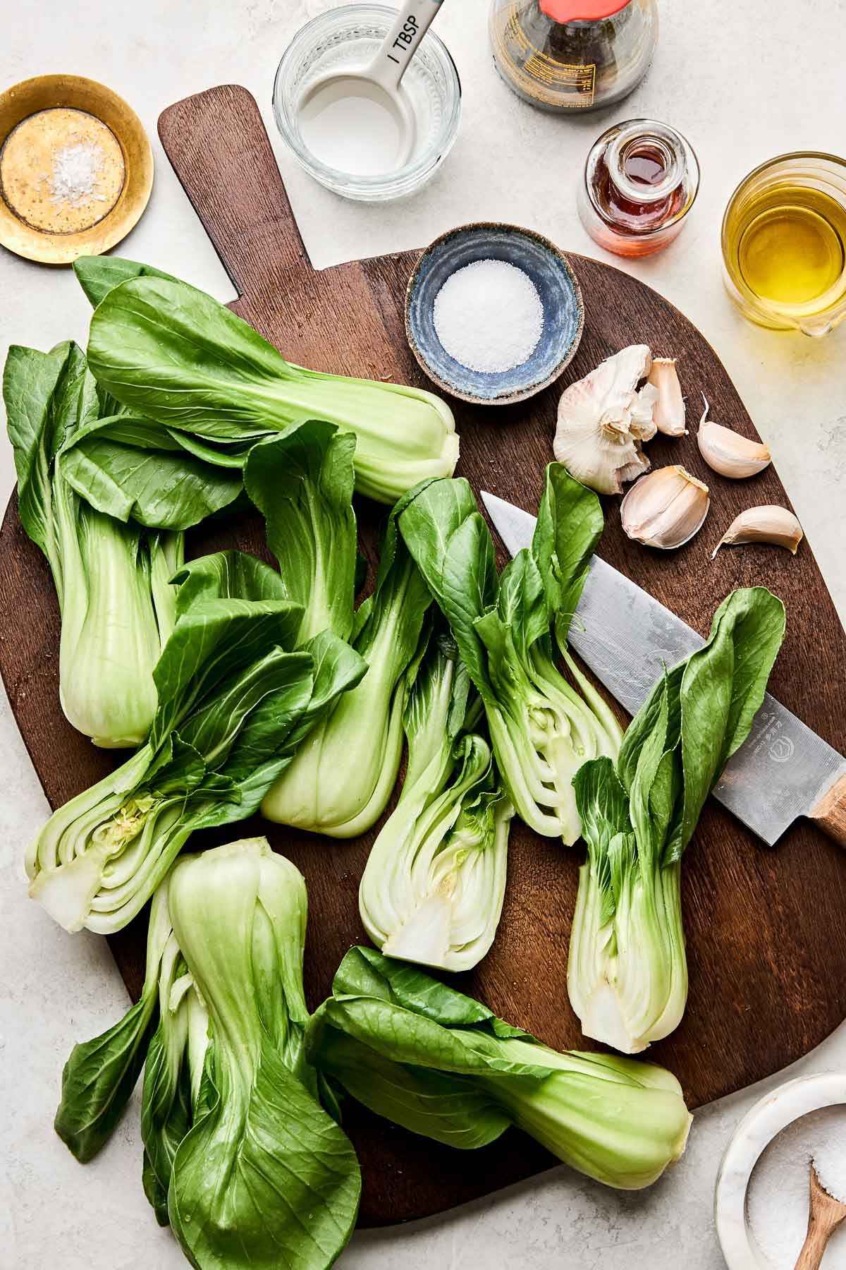 An overhead shot of ingredients displayed on a dark wood board atop a white marbled surface: baby bok choy, oil, salt, MSG, shoyu, water, toasted sesame oil, sugar, and garlic.