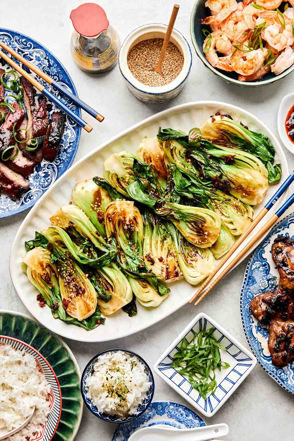 An overhead shot of sauteed baby bok choy on a white oval platter atop a white surface. Dishes of grilled meat, rice, shrimp, green onions, and sesame seeds sit alongside the platter.