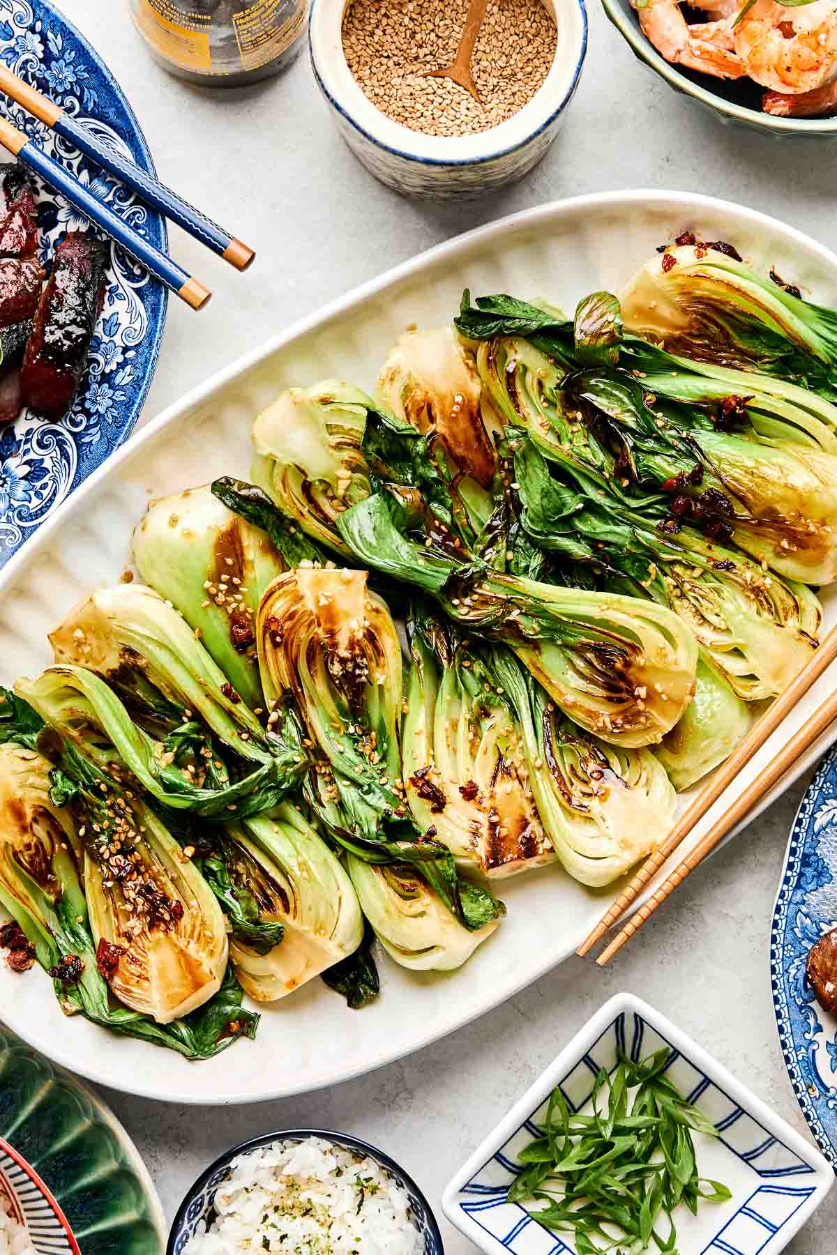 An overhead shot of sauteed baby bok choy on a white oval platter atop a white surface. Dishes of grilled meat, rice, shrimp, green onions, and sesame seeds sit alongside the platter.