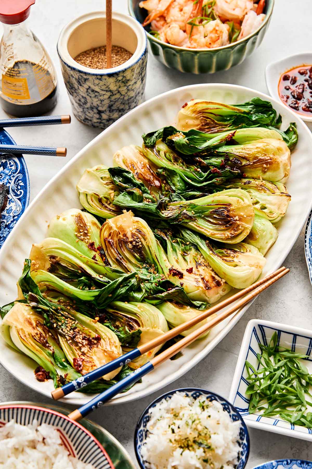 An overhead shot of steamed baby bok choy with chopsticks on a white oval platter atop a white surface. Dishes of grilled meat, rice, shrimp, green onions, and sesame seeds sit alongside the platter.