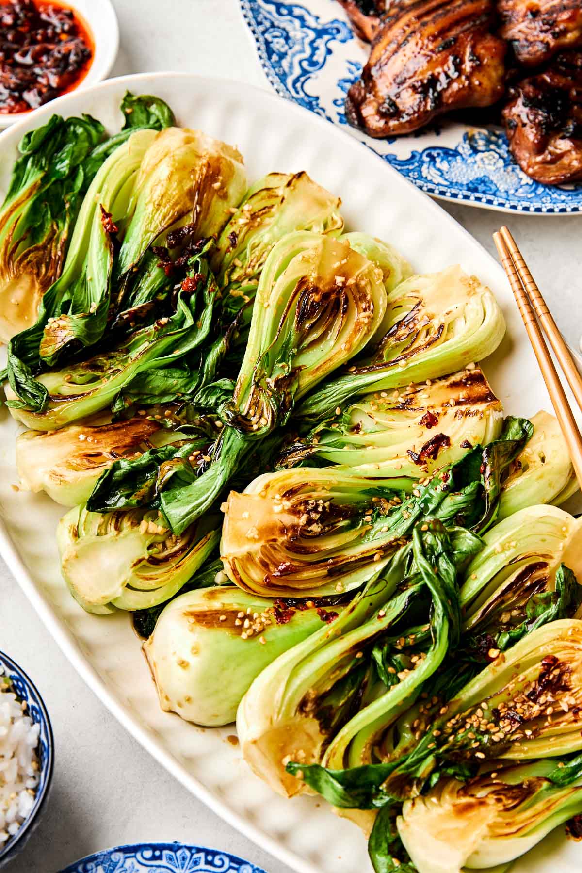 An overhead shot of steamed baby bok choy on a white oval platter atop a white surface. Dishes of grilled meat, rice and dumpling sauce sit alongside the platter.