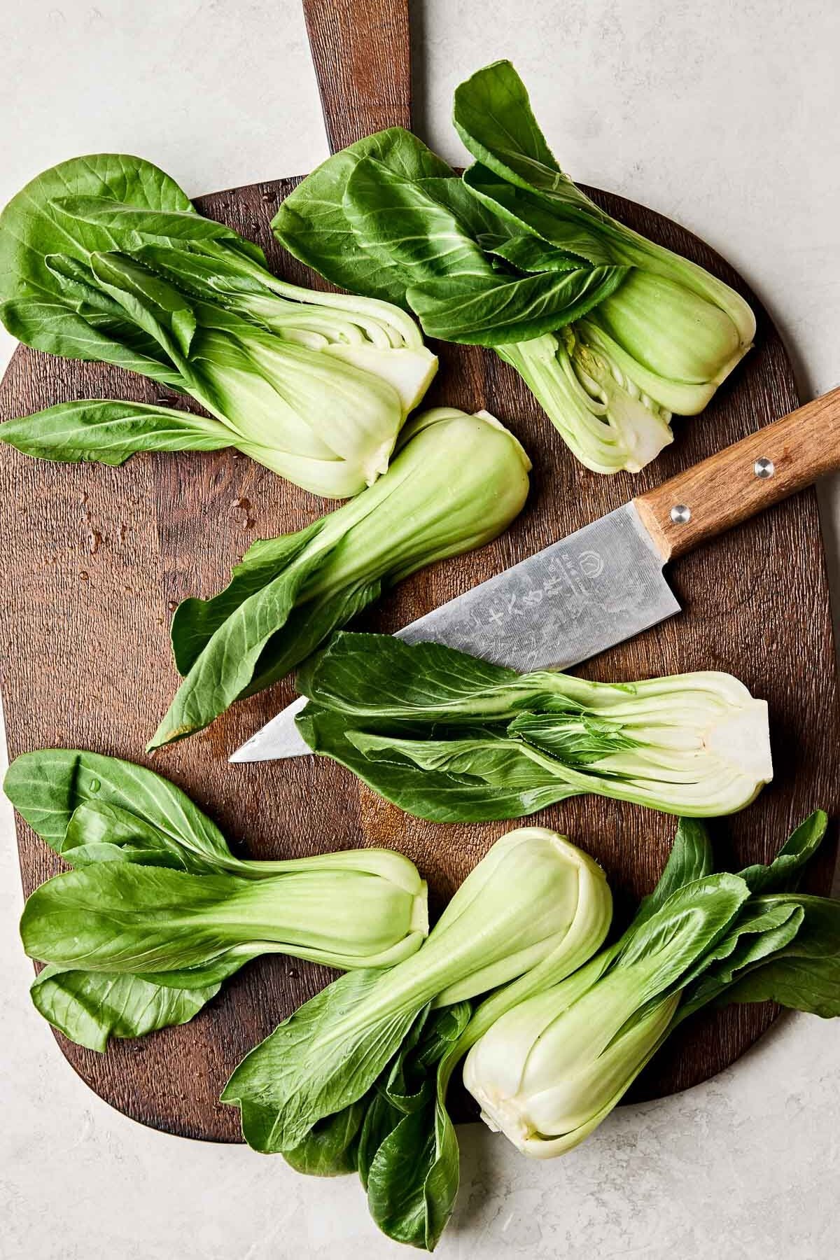 An overhead shot of baby bok choy with a chef's knife on a dark wood cutting board atop a white marbled surface.