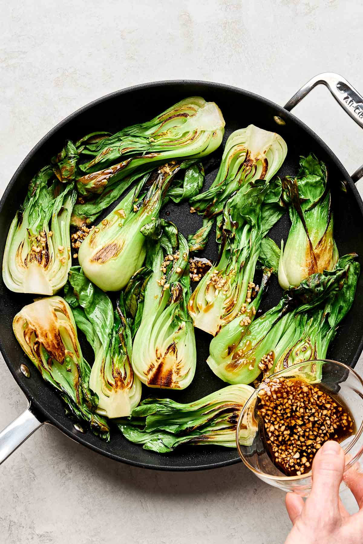 An overhead shot of a woman's hand pouring a mixture of shoyu, water, sesame oil, sugar and garlic over sauteed bok choy in a black skillet atop a white textured surface.
