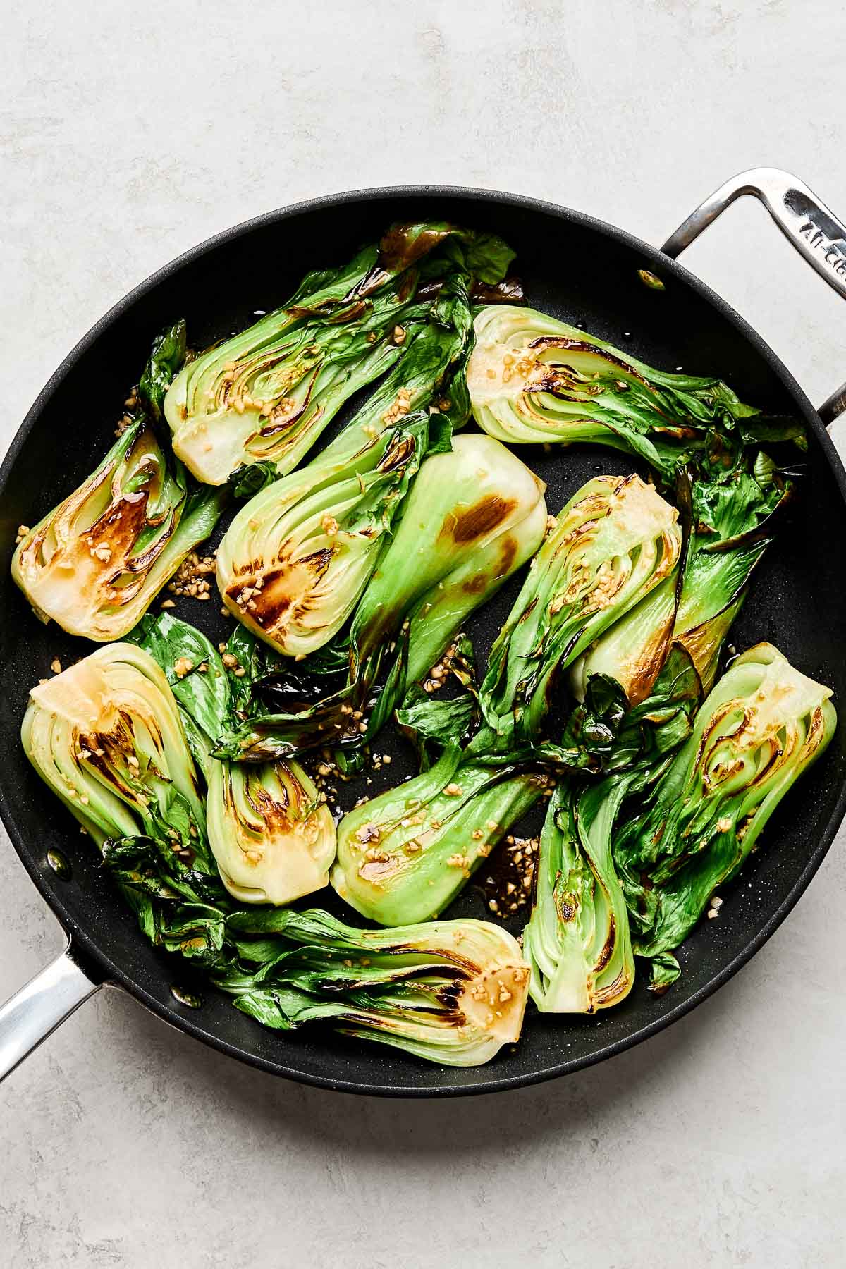 An overhead shot of sauteed baby bok choy in a black skillet atop a white textured surface.