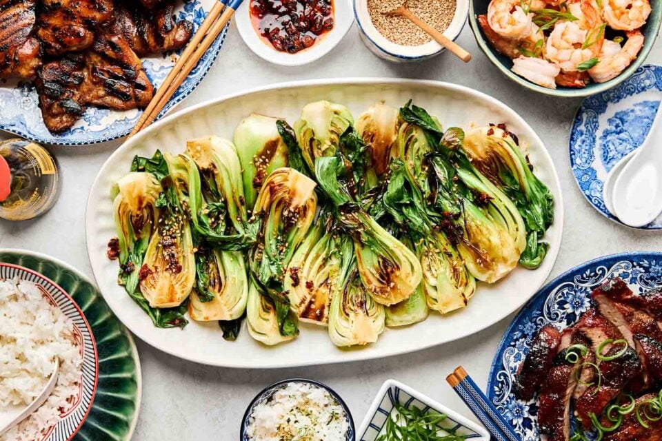 An overhead shot of sauteed baby bok choy on a white oval platter atop a white surface. Dishes of grilled meat, rice, shrimp, green onions, and sesame seeds sit alongside the platter.