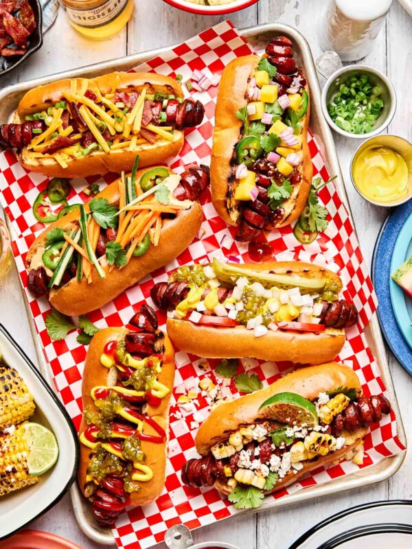 A tray of six hot dogs with spiral-cut sausages, each topped with colorful ingredients like pickled vegetables, pineapple salsa, and crumbled cheese, on red checkered paper with corn, drinks, and condiments nearby.