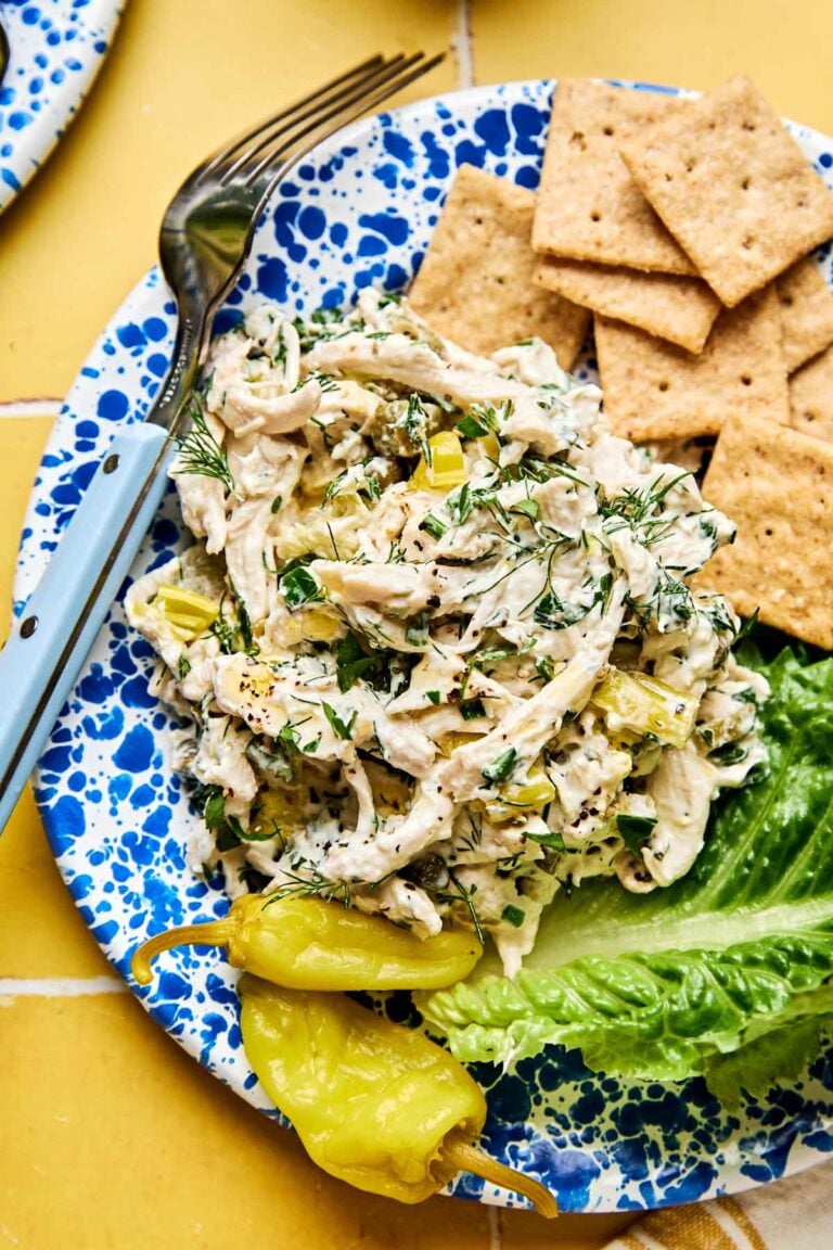A blue speckled plate with creamy chicken salad mixed with herbs and chopped celery, served with lettuce, crackers, and a yellow pepper. A fork is on the side of the plate.