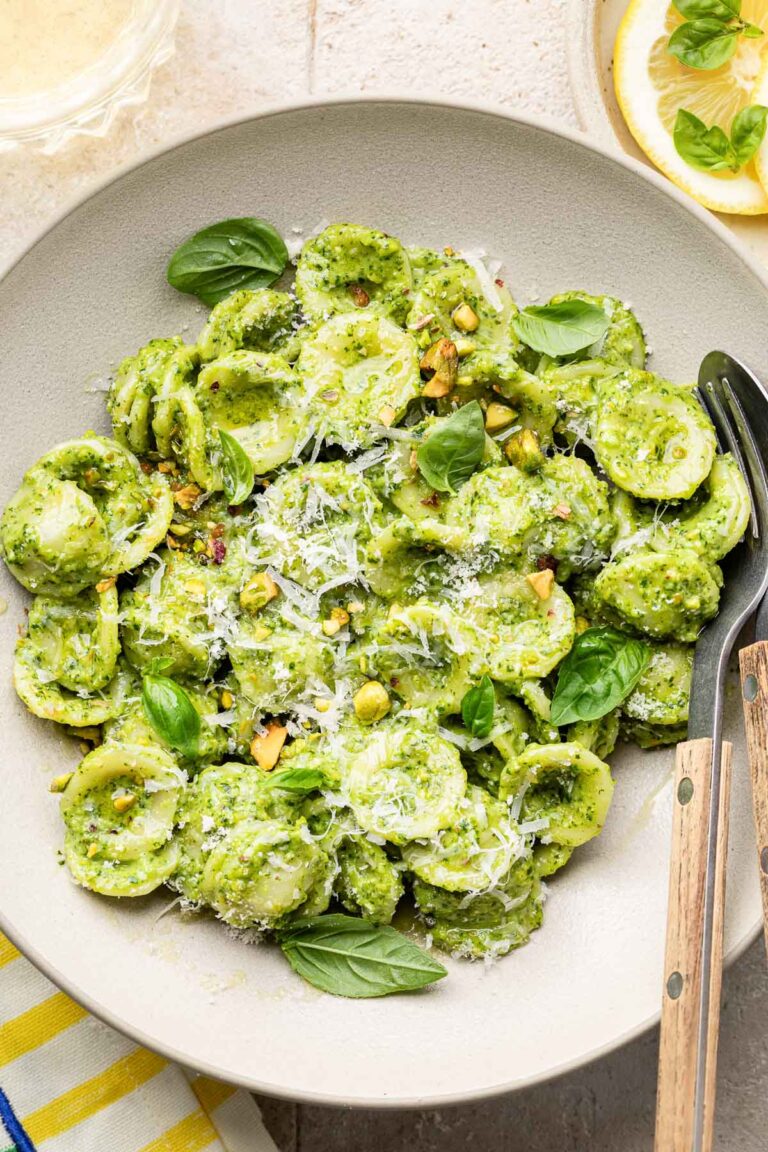 A plate of orecchiette pasta coated in green pesto sauce, topped with grated cheese, fresh basil leaves, and chopped pistachios, served with a fork and spoon on the side.