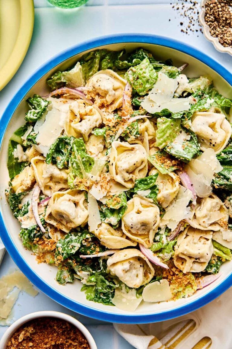 A bowl of tortellini Caesar salad with cheese tortellini, romaine lettuce, shaved parmesan, red onion, croutons, and Caesar dressing, garnished with cracked black pepper.