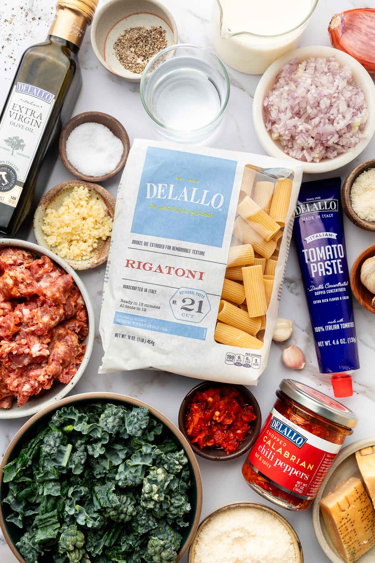 An overhead view of raw rigatoni pasta, ground sausage, tomato paste, canned chili peppers, kale, grated cheese, olive oil, minced shallots, spices, and cream arranged on a white surface.