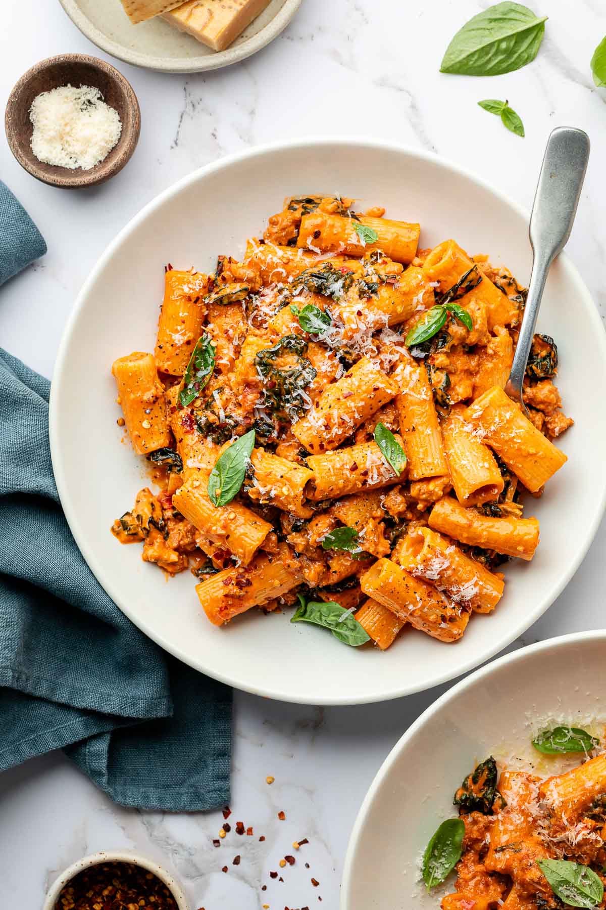 A plate of spicy rigatoni pasta in a creamy tomato sauce with kale, sprinkled with grated cheese and fresh basil leaves, served with a fork. Small bowls of grated cheese and red pepper flakes are nearby.