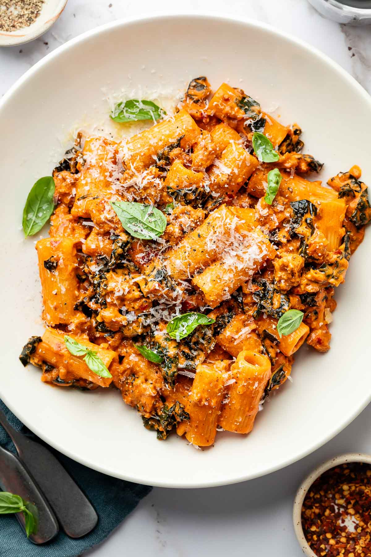 A white plate filled with rigatoni pasta in a creamy tomato sauce with ground sausage, wilted greens, fresh basil leaves, and grated Parmesan cheese, garnished with extra basil and cheese.