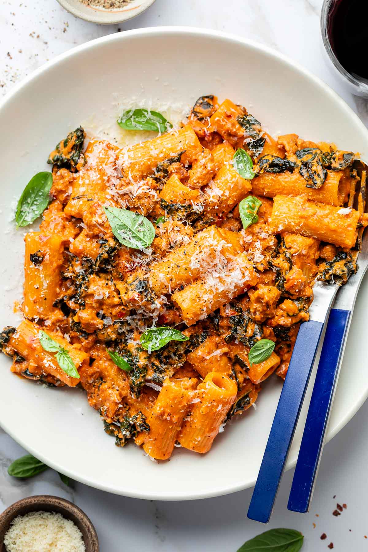A white plate of rigatoni pasta mixed with a creamy tomato sauce, ground sausage, spinach, and topped with grated cheese and fresh basil leaves. A fork and knife are placed on the side of the plate.