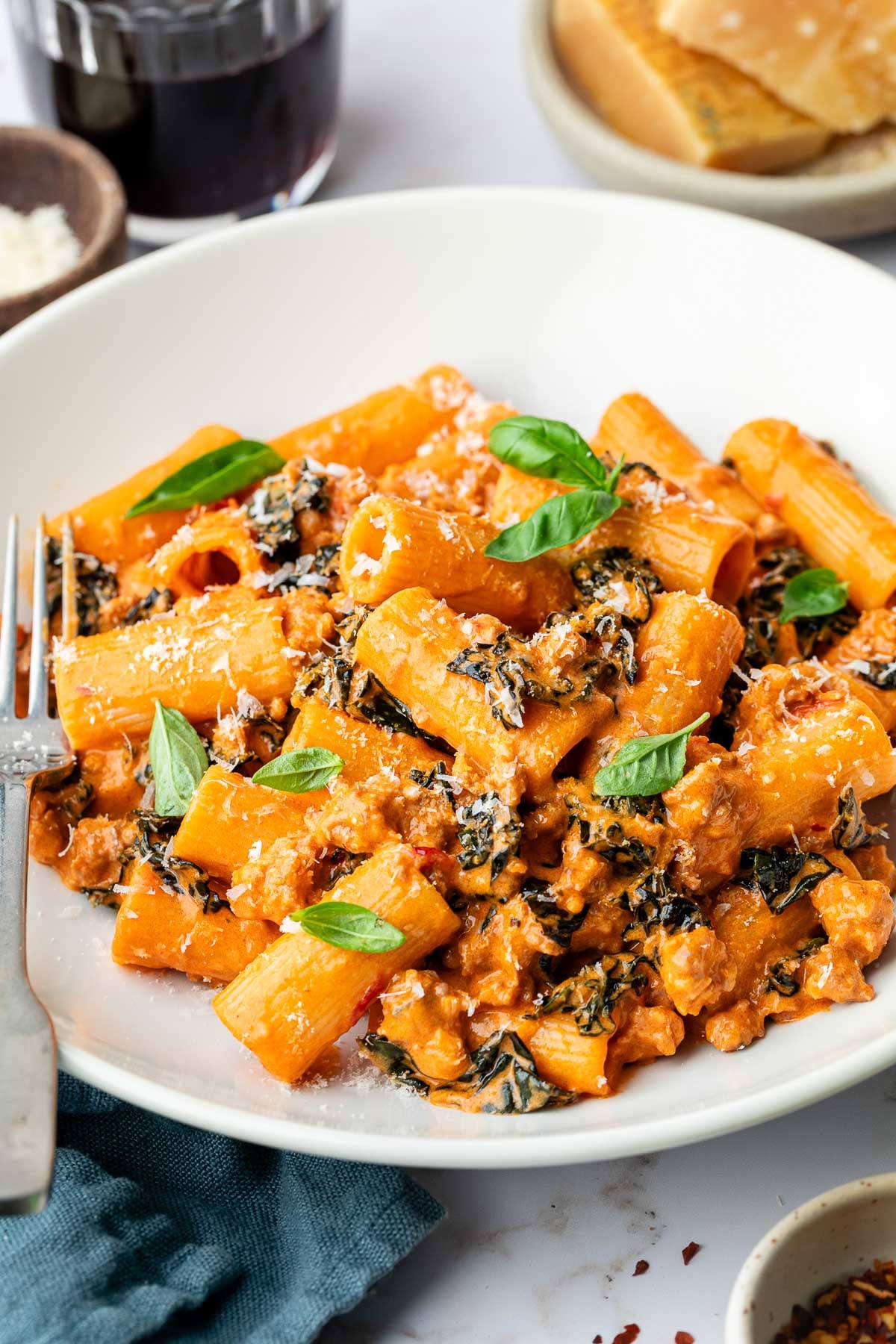 A white bowl filled with rigatoni pasta in a creamy tomato sauce with ground sausage, wilted greens, and topped with fresh basil and grated cheese. A fork, glass of red drink, bread, and napkin are nearby.
