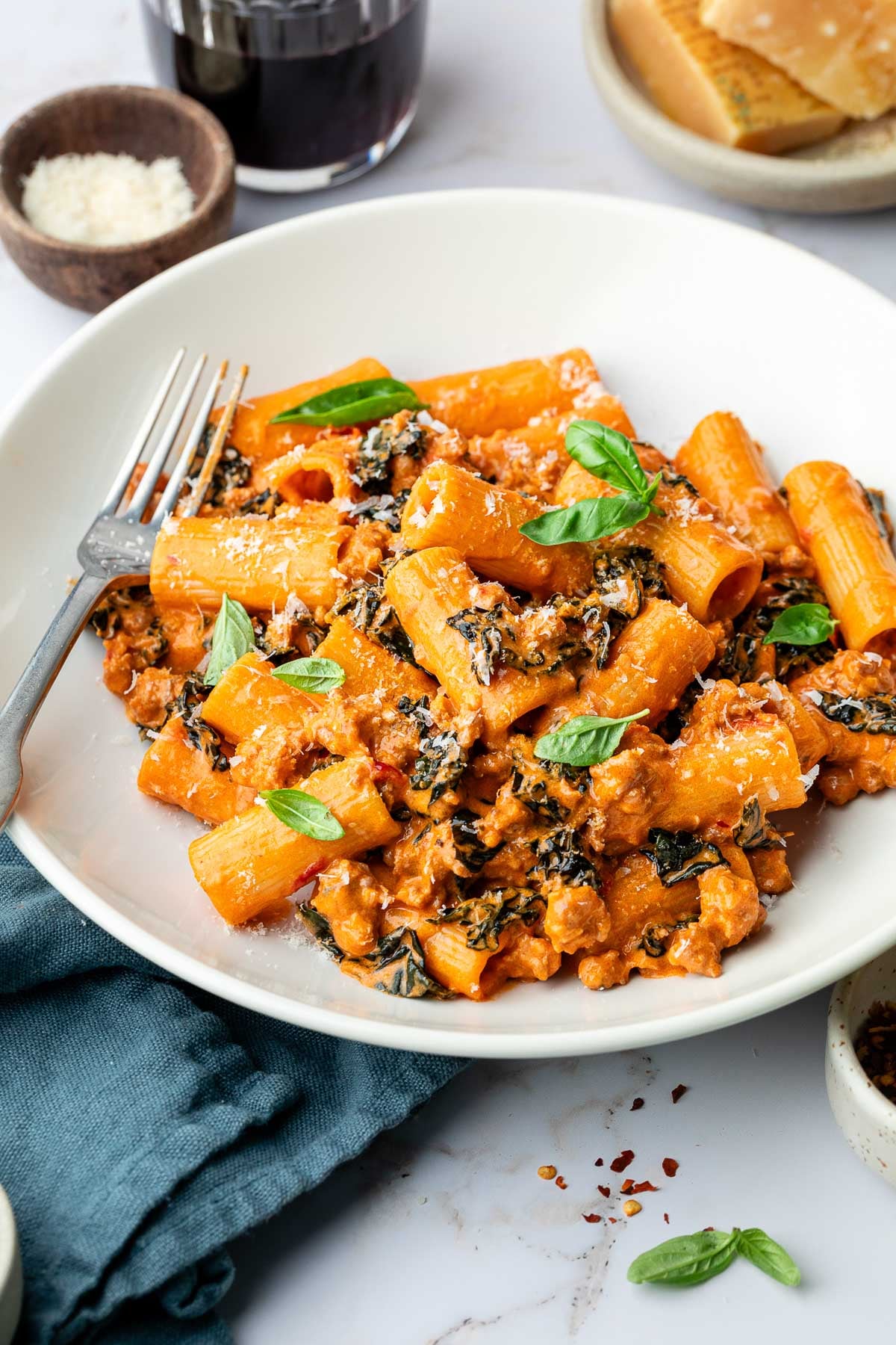 A bowl of spicy rigatoni pasta with creamy tomato sauce, ground sausage, and kale, garnished with fresh basil and grated cheese. A fork rests on the plate, with a glass of red wine and bread in the background.