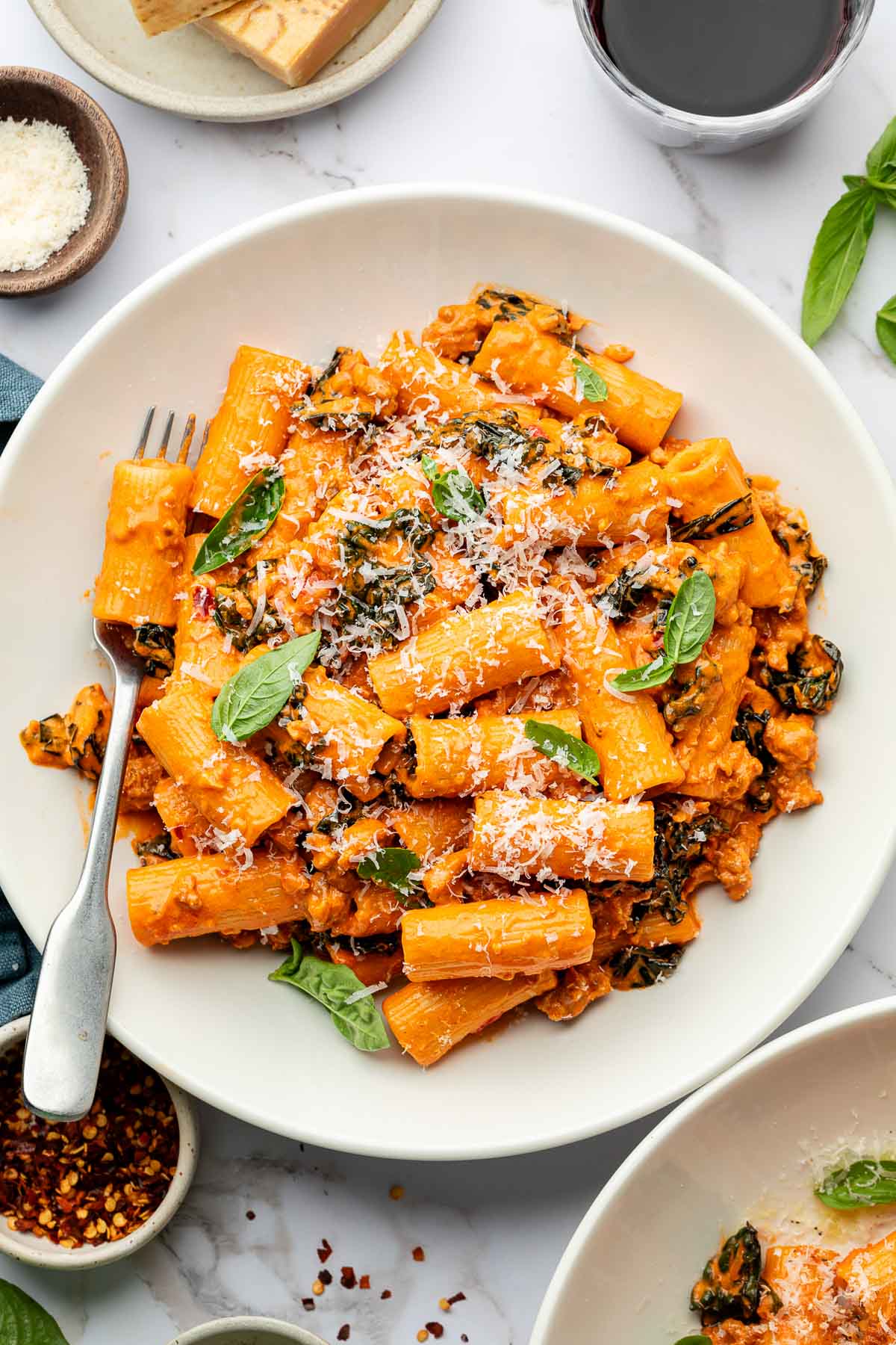 A white plate of spicy rigatoni pasta in a creamy tomato sauce with kale, topped with grated cheese and fresh basil leaves. A fork rests on the plate, and small bowls of Parmesan and chili flakes are nearby.