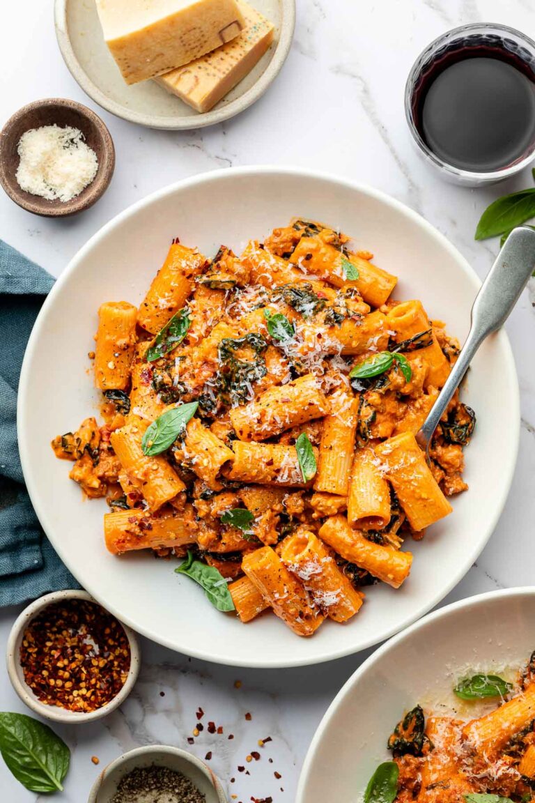 A white plate of spicy rigatoni pasta with tomato sauce, kale, sausage, and grated cheese, garnished with basil leaves. Surrounding the plate are small bowls of grated cheese, pepper, red pepper flakes, and a glass of red wine.