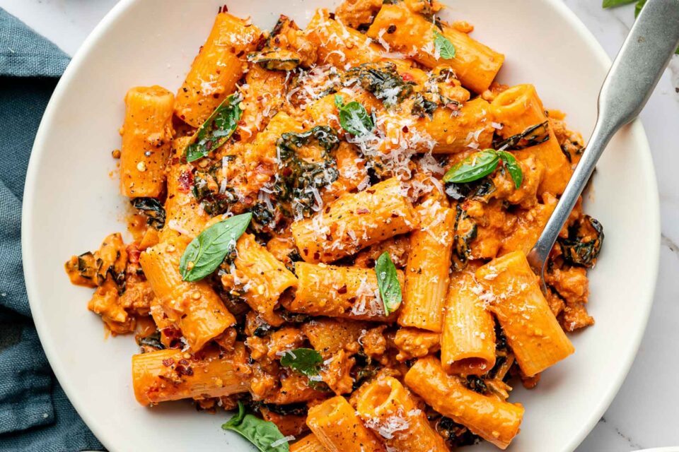 Spicy rigatoni pasta in a creamy tomato sauce with ground sausage, topped with grated cheese and fresh basil leaves, served in a white bowl with a fork.