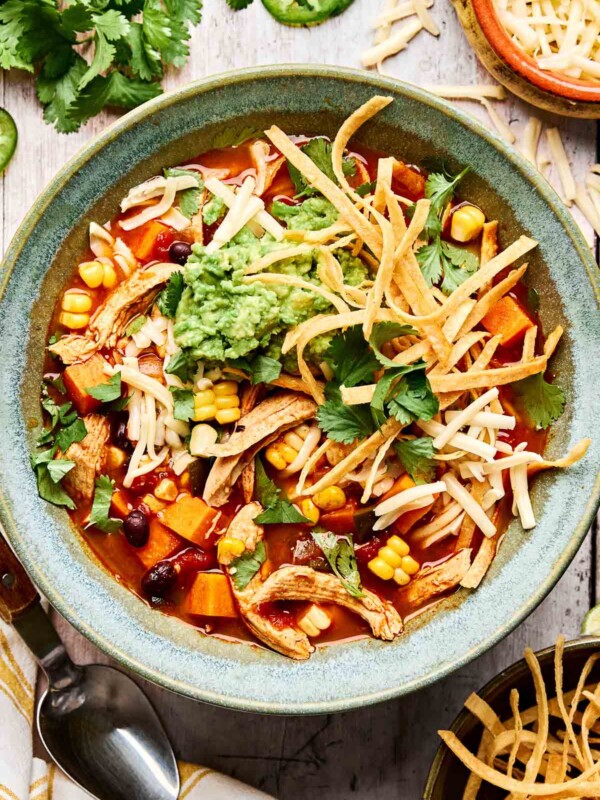 A bowl of tortilla soup topped with shredded cheese, crispy tortilla strips, guacamole, corn, cilantro, and black beans, surrounded by fresh cilantro and a bowl of extra toppings.