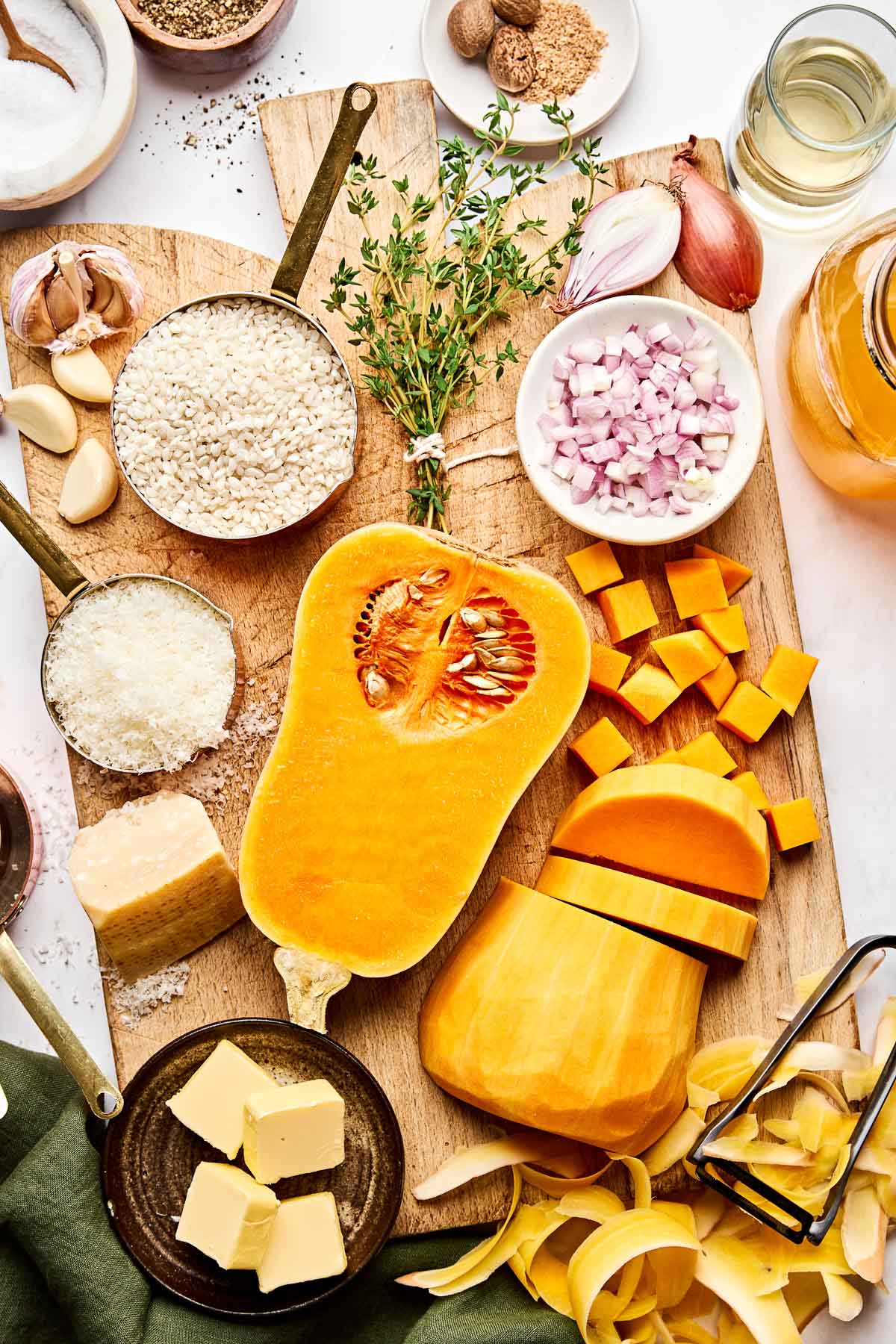 A wooden board with halved and cubed butternut squash, rice, chopped shallots, garlic, thyme, grated cheese, butter, a wedge of parmesan, seasonings, and a peeler with squash skins.