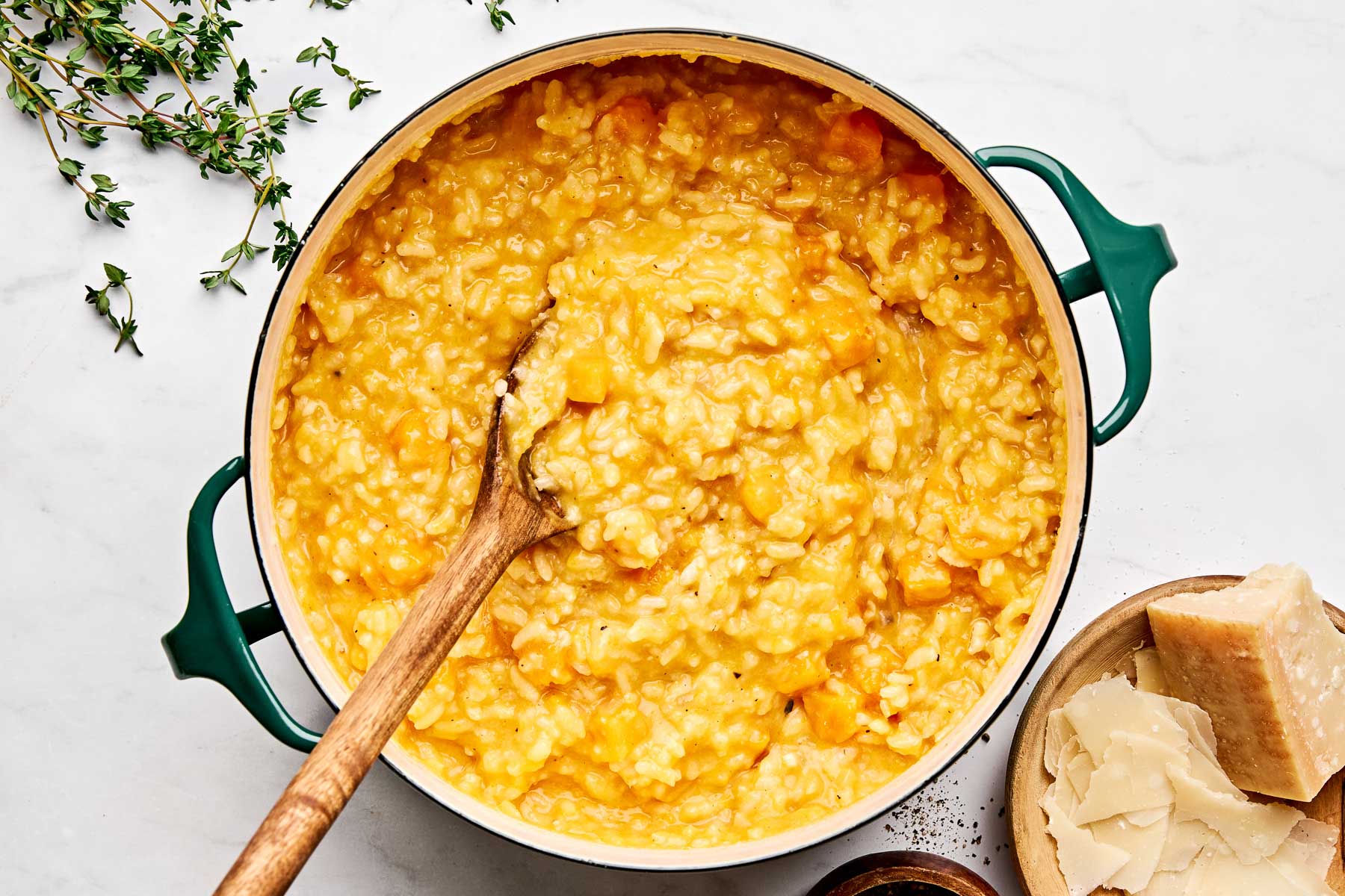 A pot of creamy risotto with orange chunks, likely butternut squash, being stirred with a wooden spoon. Fresh thyme sprigs, a bowl of Parmesan cheese shavings, and black pepper are nearby on a white surface.