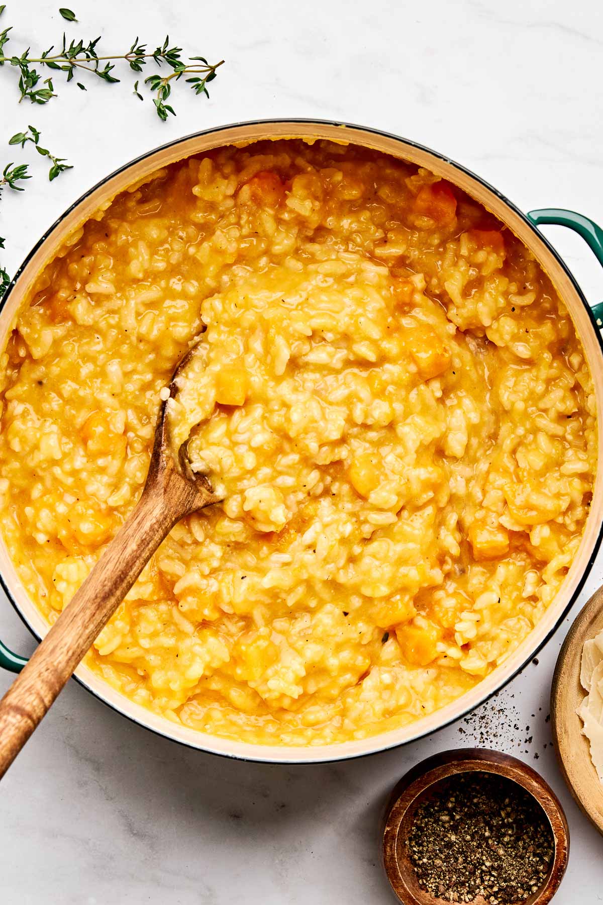 A pot of creamy butternut squash risotto with a wooden spoon, surrounded by fresh thyme and a bowl of cracked black pepper on a white marble surface.