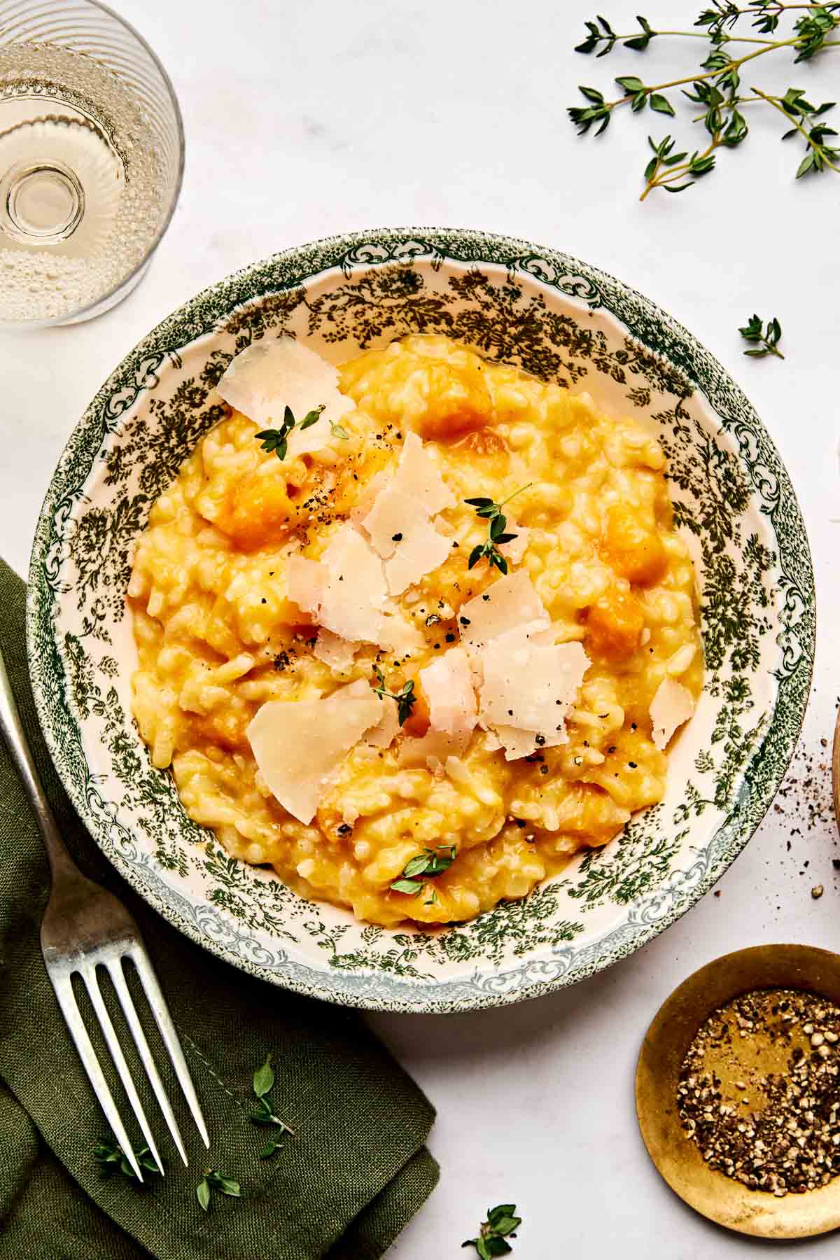 A bowl of creamy butternut squash risotto topped with shaved cheese, black pepper, and fresh thyme, set on a patterned plate. A fork, napkin, glass of white wine, pepper, and herb sprigs are nearby on a white surface.