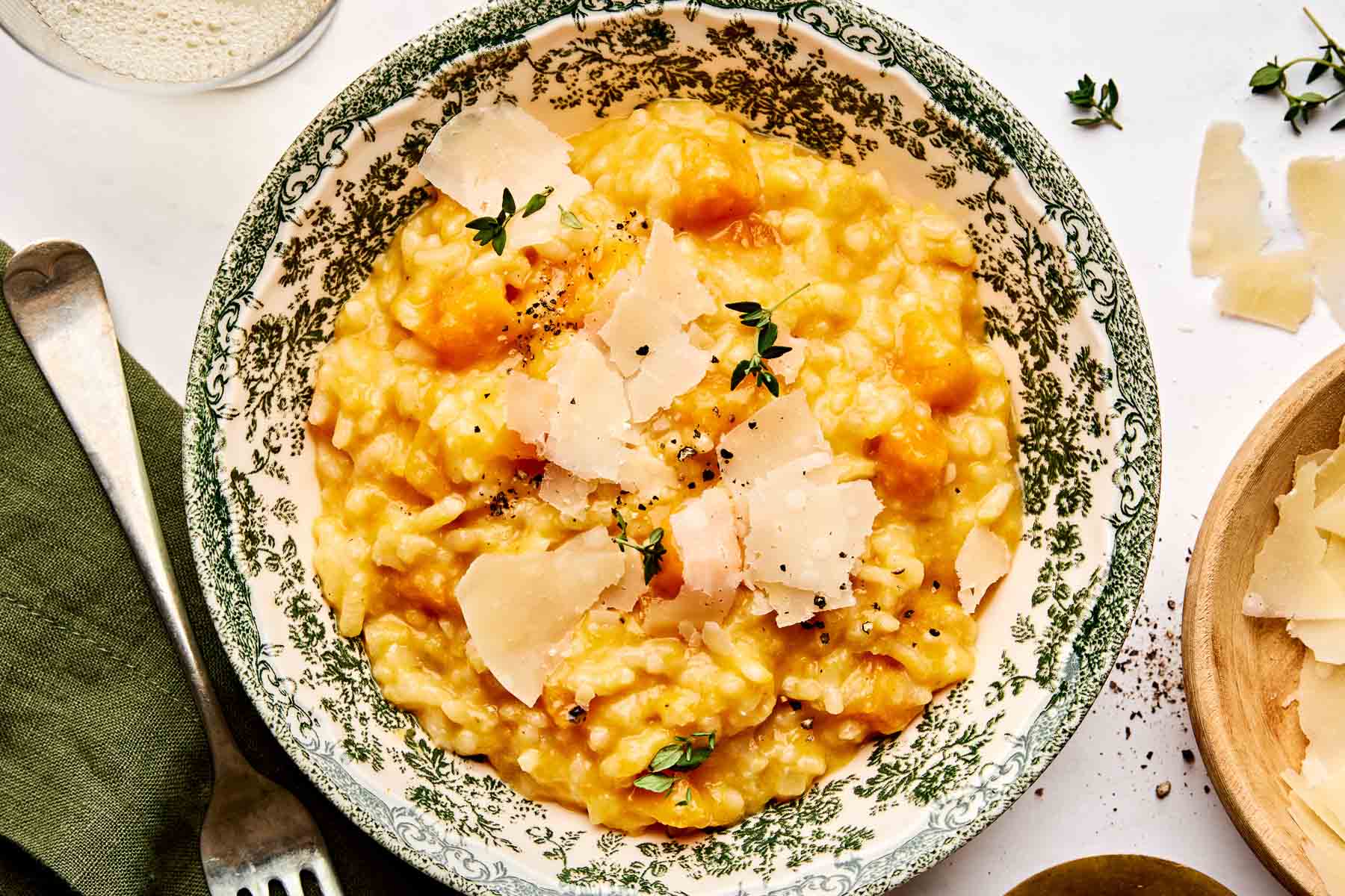 A bowl of creamy risotto with chunks of butternut squash, topped with shaved parmesan, black pepper, and fresh thyme, served in a patterned dish. A fork and napkin are on the side.