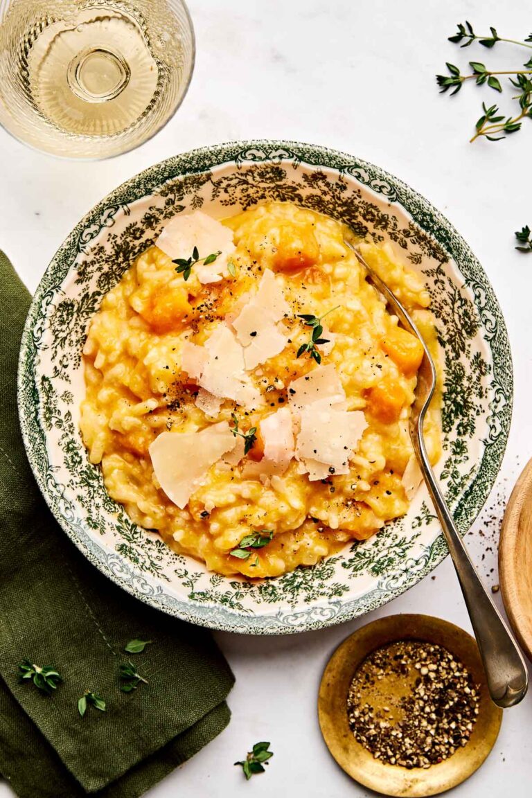 A bowl of creamy baked butternut risotto topped with shaved parmesan and fresh herbs sits on a green-patterned plate, next to a green napkin, a glass of white wine, and a small dish of cracked black pepper.