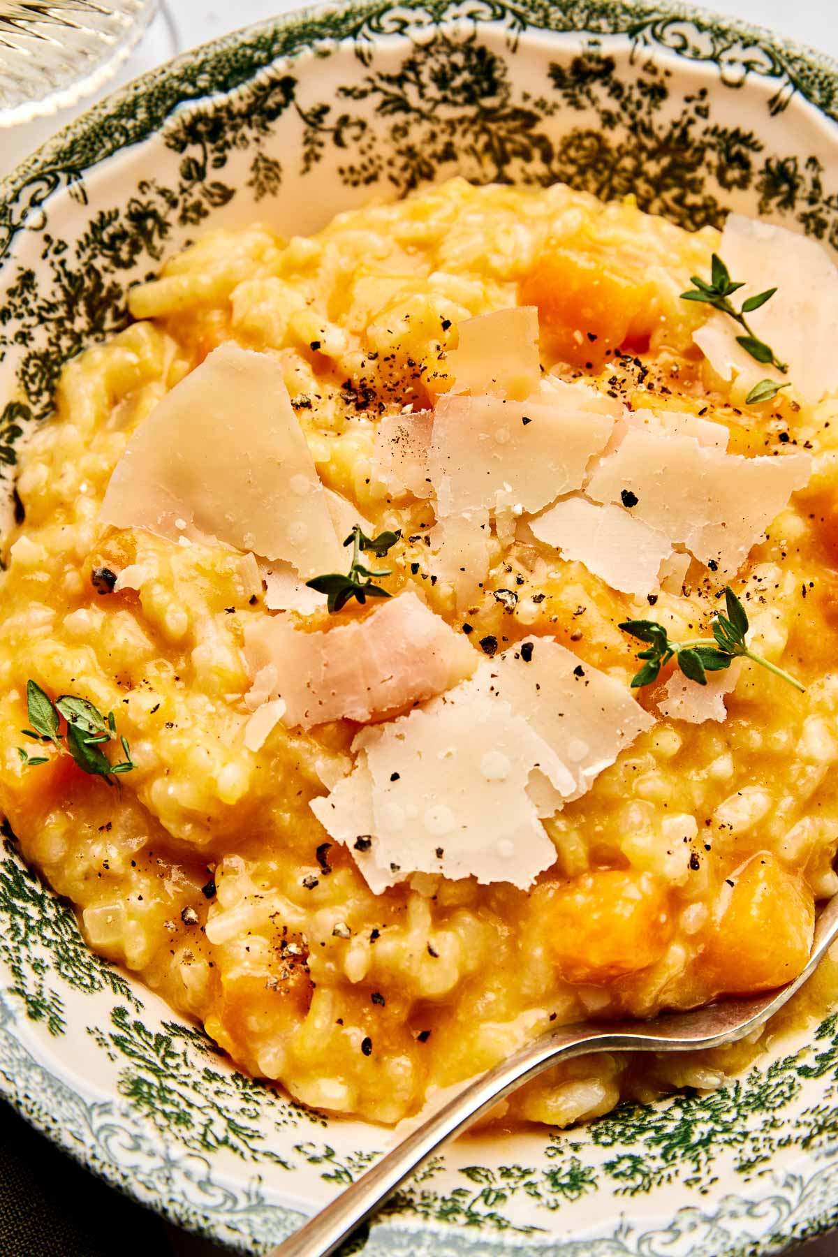 A close-up of creamy risotto with chunks of butternut squash, topped with shaved Parmesan cheese, black pepper, and fresh thyme, served in a decorative bowl with a spoon.