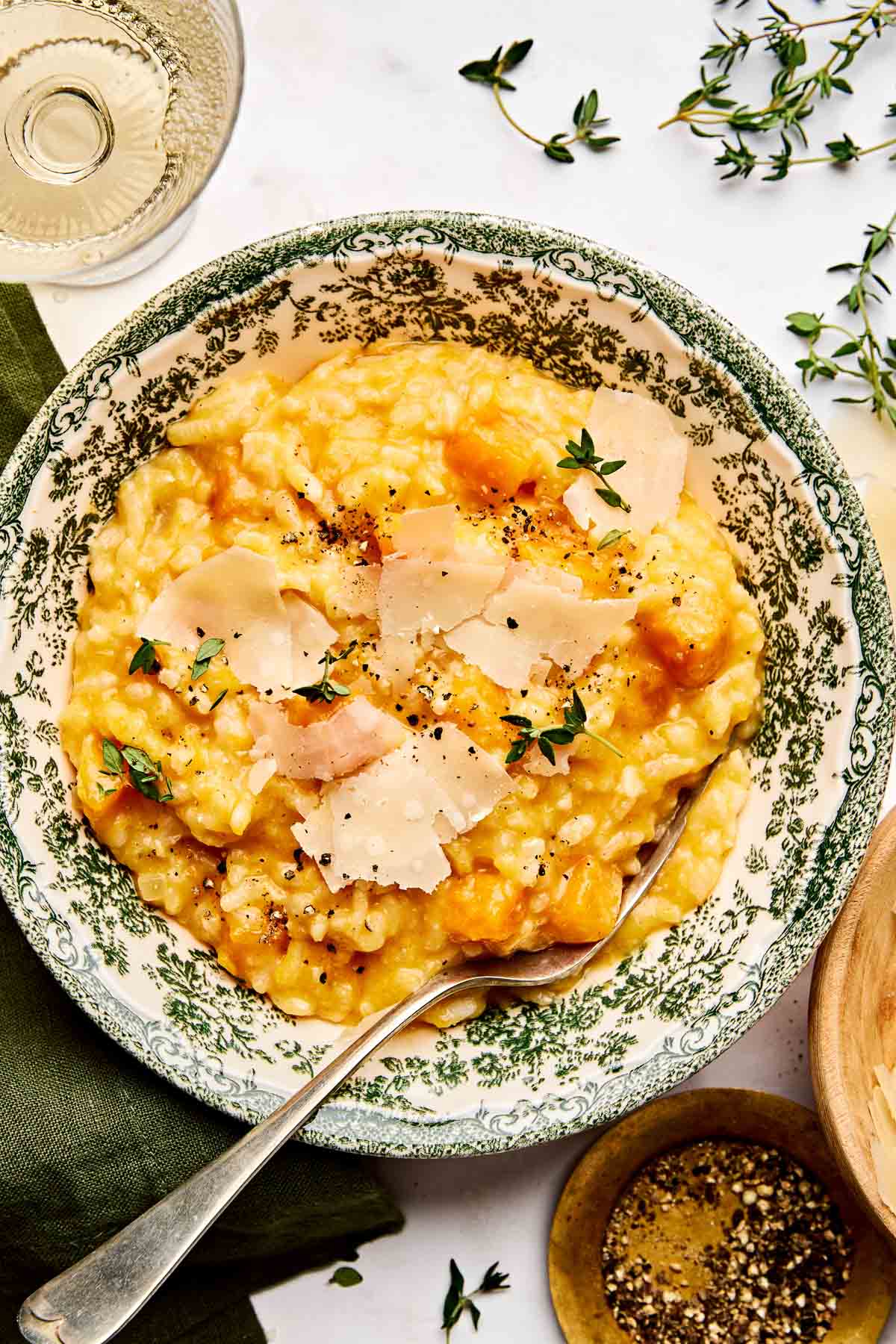 A bowl of creamy butternut squash risotto topped with shaved parmesan, black pepper, and fresh thyme. A spoon rests in the bowl, and side items like a glass of wine and a small dish of pepper are nearby.