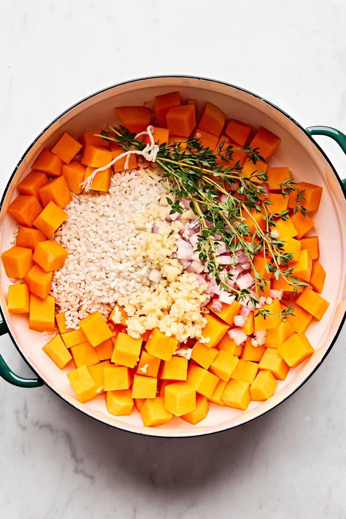 A pot filled with chopped butternut squash, arborio rice, minced garlic, diced shallots, and sprigs of fresh thyme, arranged in sections on a white surface.