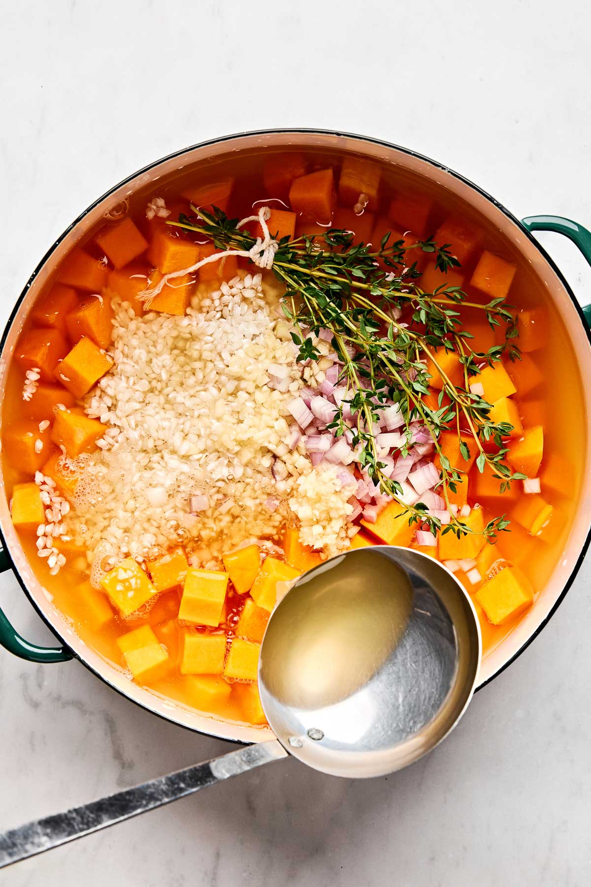 A pot filled with cubed butternut squash, chopped onions, garlic, fresh thyme sprigs, and uncooked barley in broth, with a ladle resting on the side.