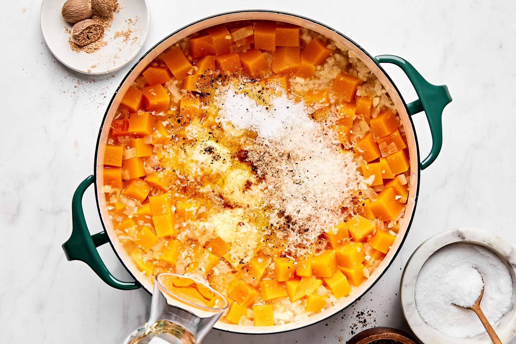 A green-handled pot filled with cubed butternut squash, diced onions, shredded cheese, seasoning, and grated nutmeg, with a measuring cup pouring liquid in. Bowls of salt and nutmeg are nearby on a marble surface.