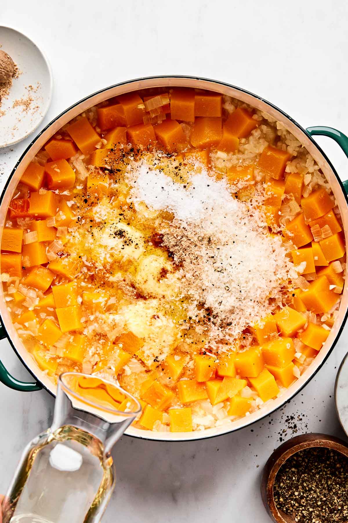 A large pot filled with cubed butternut squash, chopped onions, grated cheese, spices, and seasonings, with broth being poured in, prepared on a white countertop with small bowls of spices nearby.