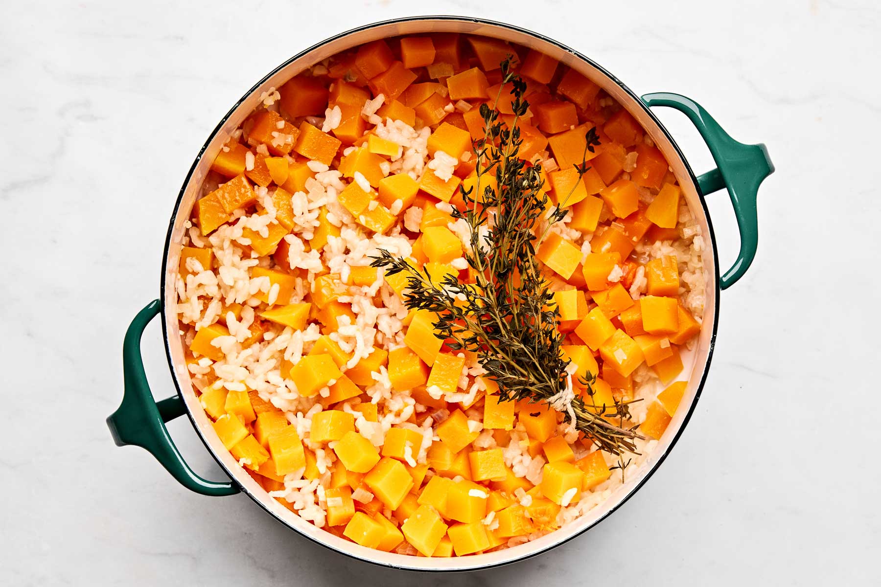 A pot of risotto with diced butternut squash and sprigs of fresh thyme on top, seen from above, on a white surface.