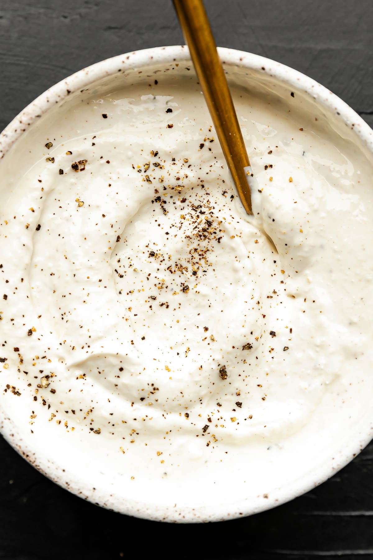 A bowl of creamy horseradish sauce topped with cracked black pepper, with a gold spoon resting inside, on a dark background.