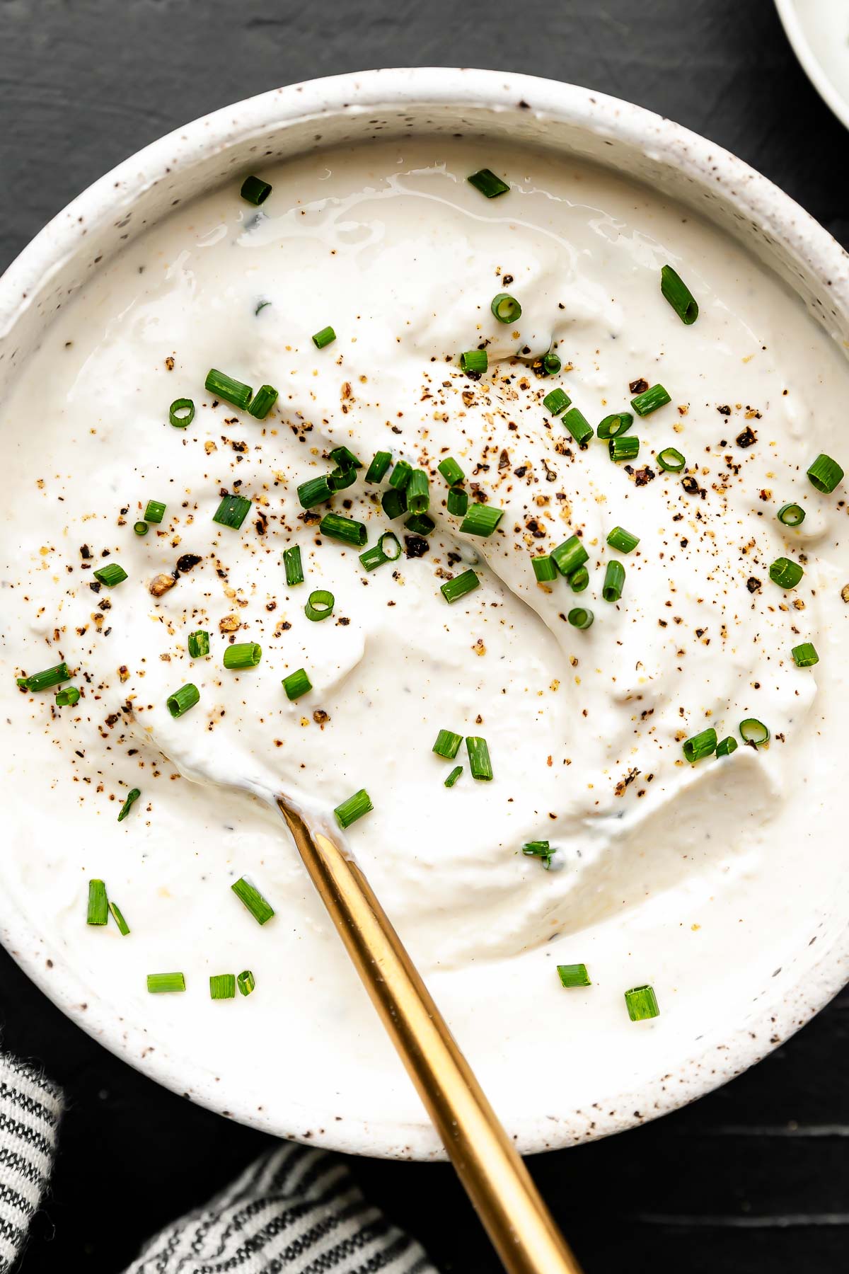 A bowl of creamy white horseradish sauce topped with chopped chives and cracked black pepper, with a gold spoon resting inside. The bowl is placed on a dark surface.