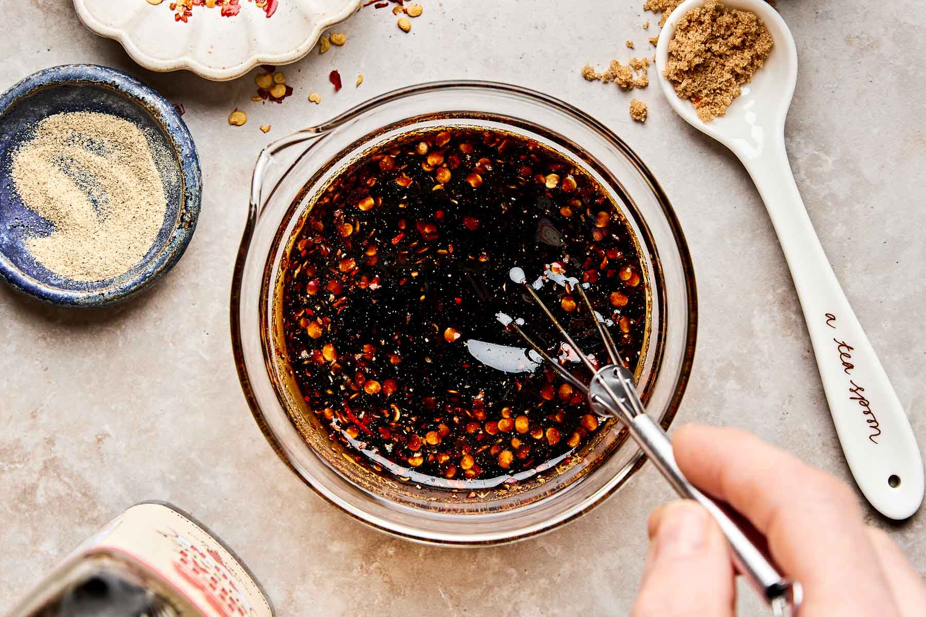 A hand uses a small whisk to mix a dark, speckled sauce in a glass bowl. Surrounding the bowl are a spoon of brown sugar, a small dish of pale seasoning, and a plate with red pepper flakes.
